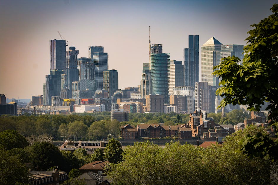 A cityscape shot of Greenwich's historic white buildings with domed towers in the foreground, surrounded by lush green trees. Beyond the historic structures, a modern skyline of high-rise office build