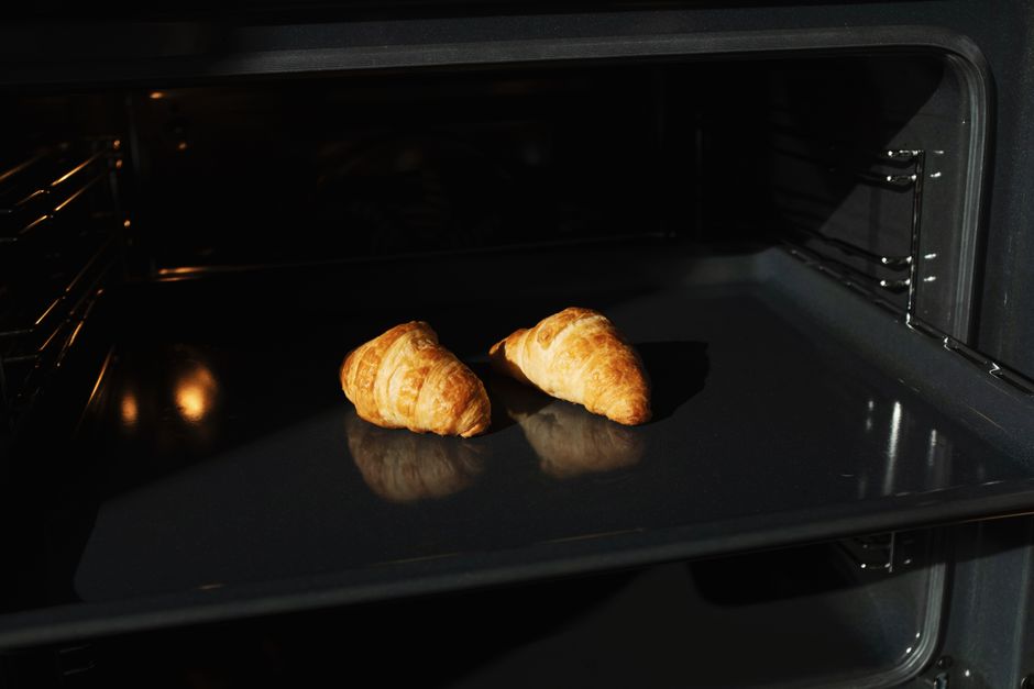 Two golden-brown croissants placed on a black baking tray inside a modern oven with stainless steel interior, illuminated by oven light. The oven's interior walls appear clean, and the baking tray is 
