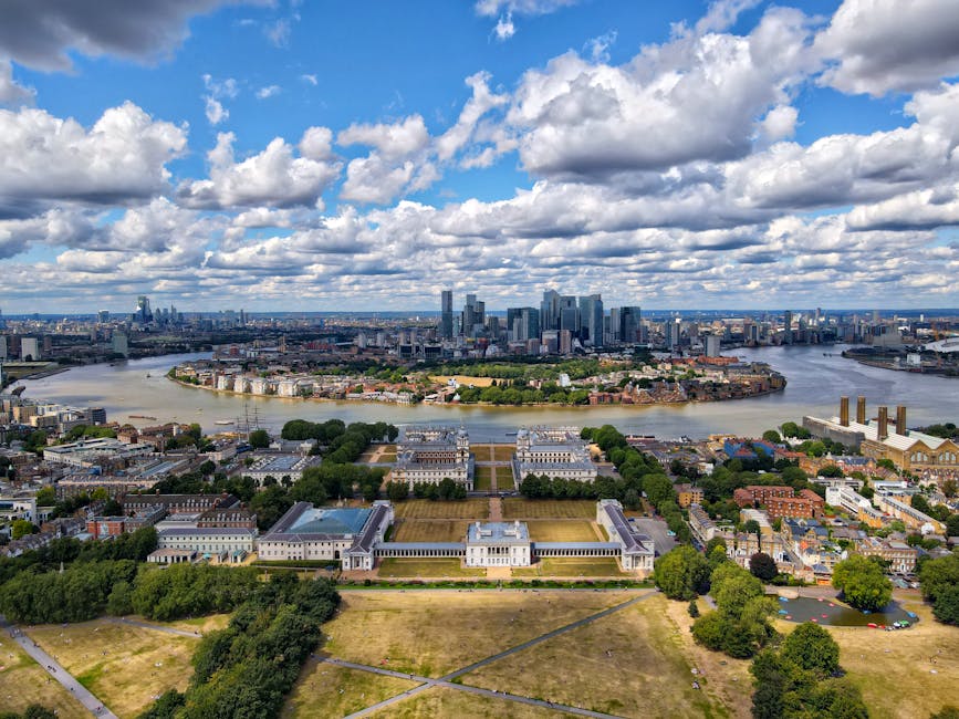 A panoramic view of a modern city skyline with tall glass skyscrapers reflecting sunlight, set against a partly cloudy blue sky. In the foreground, a lush green park with a variety of mature trees and