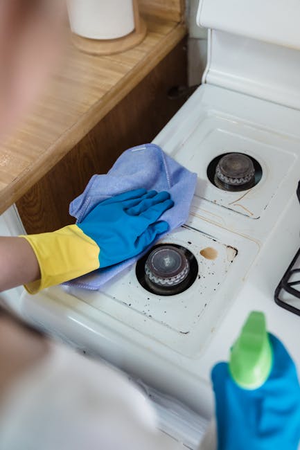 A woman in a wheelchair is cleaning a kitchen countertop with a blue cloth, holding a spray bottle of cleaning solution in her right hand. The kitchen features a white marble backsplash, stainless ste